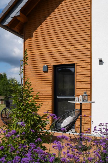 Detailaufnahme der Fassade aus Fichtenholz-Rhombusprofil am Einfamilienhaus Lichtenau mit schwarzem Fensterrahmen, Dekotisch und violetten Blumen im Vordergrund